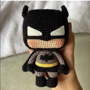May include: A small, handmade crochet doll of a superhero character featuring a black cowl with pointed ears, white felt eyes, and a beige face with pink cheeks. The body is grey with a black chest emblem, a yellow belt, and black limbs.