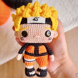 May include: A crochet doll featuring bright yellow spiky hair, a white headband with a black spiral symbol, large black circular eyes, and three small horizontal lines on each cheek. The doll wears an orange and black outfit with black shoes.