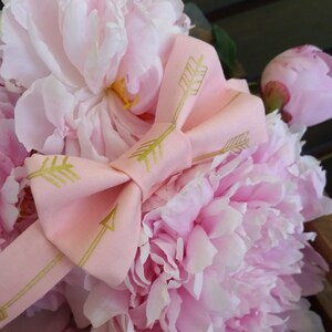May include: A bouquet of pink peonies tied with a pink ribbon with gold arrow designs.