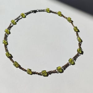 Könnte beinhalten: Eine zarte Halskette mit kleinen, quadratischen, hellgrünen Edelsteinen, die auf einer dünnen, bronzefarbenen Kette aufgereiht sind. Die Edelsteine sind gleichmäßig um die Halskette verteilt und erzeugen ein schlichtes, aber elegantes Design. Die Halskette wird auf einer weißen Oberfläche präsentiert.