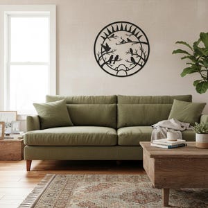 May include: A black metal wall decoration featuring birds on branches inside a circular frame. It's mounted on a neutral wall above a green sofa. The room includes a wooden coffee table and a patterned rug. The overall style is modern.