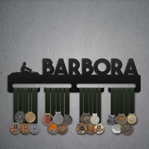 May include: Black metal medal display with the name "BARBORA" cut out. The display features a silhouette of a rower and holds several medals with green ribbons. The medals are of various colors and designs.