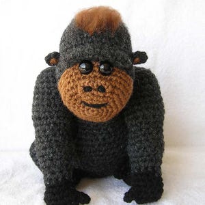 Può includere: Un giocattolo di gorilla all'uncinetto fatto a mano. Il gorilla è principalmente grigio scuro con un viso marrone e un ciuffo di capelli castani. Ha grandi occhi neri ed è seduto in posizione eretta.