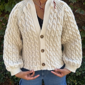 May include: Cream-colored cable knit cardigan with a V-neck and three brown buttons. The sweater has a textured, woven pattern and long sleeves. The cardigan is a classic style, suitable for layering.