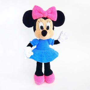 Puede incluir: Mu&ntilde;eca de ganchillo hecha a mano de un personaje de dibujos animados. La mu&ntilde;eca tiene orejas negras, cara color canela y un gran lazo rosa. Viste un vestido azul, guantes blancos y zapatos rosas. La mu&ntilde;eca est&aacute; sobre un fondo blanco.