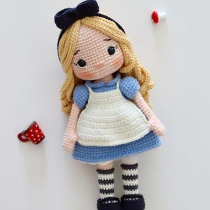 Può includere: Bambola all'uncinetto raffigurante un personaggio con capelli biondi, un vestito blu e un grembiule bianco. La bambola ha calzini a righe bianche e nere e un fiocco blu scuro. È visibile una piccola tazza rossa a pois bianchi.