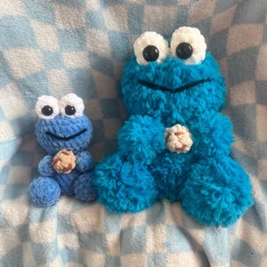 May include: Two blue crochet Cookie Monster plush toys. The larger one has a cookie in the center. Both have white eyes with black pupils and a smiling mouth. The toys are on a blue and white checkered blanket.