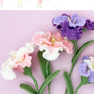 Può includere: Fiori all'uncinetto fatti a mano nei toni del bianco, rosa e viola, con steli e foglie verdi. I fiori sono disposti su uno sfondo lilla chiaro. I fiori hanno centri gialli e bordi dei petali dettagliati.