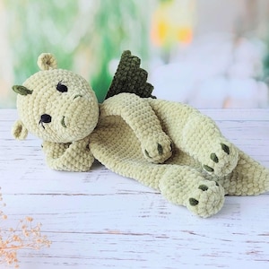 May include: A handmade, light green crocheted dragon plush toy. The dragon has black eyes, a smiling mouth, and dark green accents on its wings and feet. The plush is lying down on a white wooden surface.