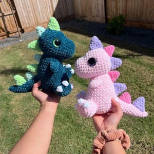 May include: Two handmade crochet dinosaur toys. One is teal with green accents, the other is pink with purple and pink accents. Both have large black eyes and are held up by hands.