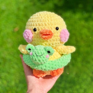 Puede incluir: Un juguete de ganchillo hecho a mano de un patito amarillo, naranja y rosa, con una rana verde envuelta alrededor. El patito tiene ojos negros y un pico naranja. La rana tiene mejillas rosas y una cara sonriente.