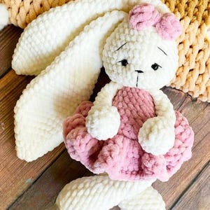 Può includere: Una bambola coniglietto all'uncinetto bianca e rosa fatta a mano. Il coniglietto ha lunghe orecchie flosce, un fiocco rosa e un vestito rosa. La bambola è realizzata in filato morbido e strutturato ed è appoggiata su una superficie di legno.