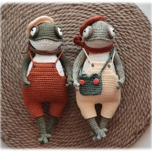 May include: Two handmade crochet frog dolls. One frog wears orange overalls, a tan beret, and a brown scarf. The other frog wears tan overalls, a red beret, and a red scarf, with a green bag. Both frogs have large eyes and are holding hands.