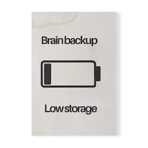 Op de afbeelding: Een notitieboek met een lichtbeige omslag. De tekst "Brain backup" staat boven een zwart batterij-icoon, met "Low storage" eronder. Het notitieboek heeft een vlek in de linkerbovenhoek.