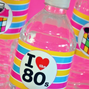 May include: Clear plastic water bottles with vibrant 80s-themed designs. The labels display "I ❤️ the 80s" and a graphic of a boombox, Rubik's cube, and cassette tape. The bottles contain pink liquid.