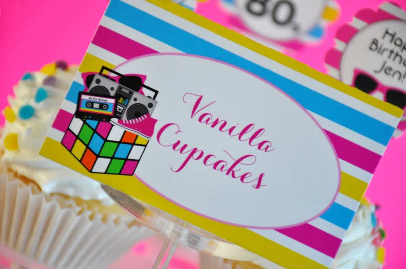 May include: A cupcake with white frosting and colorful sprinkles, topped with a decorative sign. The sign features a retro 80s theme with a boombox, cassette tape, and Rubik's Cube, and the words "Vanilla Cupcakes".