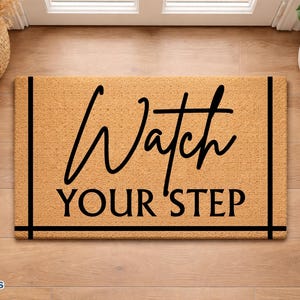 May include: A rectangular doormat with the words "Watch YOUR STEP" in black script. The mat is a light brown color with a black border. The mat is placed on a wooden floor.