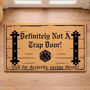 May include: A rectangular tan coir doormat featuring black printed graphics of two vertical iron-style hinges and a twenty-sided die. The text reads: Definitely Not A Trap Door! (roll for dexterity saving throw) in a bold, black gothic font.
