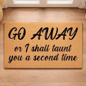 May include: A rectangular tan coir doormat featuring black text that reads GO AWAY or I shall taunt you a second time. The mat has a coarse, fibrous texture and is positioned on a light brown wooden floor surface with a plant and shoes nearby.