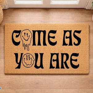 May include: A rectangular doormat with the text "COME AS YOU ARE" in black, stylized font. The mat is a natural tan color and features two smiley face designs. The mat is on a wooden floor.
