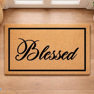 May include: A rectangular coir doormat with a black border and the word "Blessed" in elegant black script. The mat is placed on a light wood floor, near a door and a potted plant.