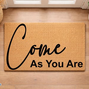 May include: A rectangular doormat with the words "Come As You Are" in black script. The mat is a light brown color and sits on a wooden floor. The scene includes a basket of dried plants, a potted plant, and a pair of shoes.
