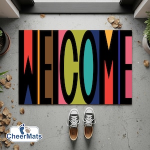 May include: A rectangular welcome mat with the word "WELCOME" in large, colorful letters. The letters are black with vertical stripes of red, orange, brown, green, teal, and pink. Two pairs of sneakers are at the bottom.