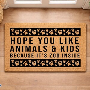 May include: A rectangular doormat with the text "HOPE YOU LIKE ANIMALS & KIDS BECAUSE IT'S ZOO INSIDE" in black. The mat has a border of black paw prints. The mat is made of a light brown material.