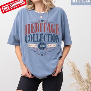 May include: A pale blue t-shirt with the words "HERITAGE COLLECTION" in red and navy blue. The shirt also features the text "EST. 1978" and the words "QUALITY", "TIMELESS", "AUTHENTIC", "ORIGINAL", and "ENDURING".