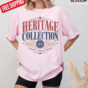 May include: A pale pink t-shirt featuring the words "HERITAGE COLLECTION" in a retro style. The shirt has the text "EST. 1978" and the words "QUALITY", "TIMELESS", "AUTHENTIC", "ORIGINAL", and "ENDURING".