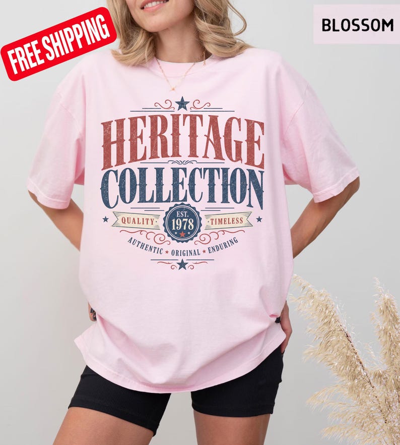 May include: A pale pink t-shirt featuring the words "HERITAGE COLLECTION" in a retro style. The shirt has the text "EST. 1978" and the words "QUALITY", "TIMELESS", "AUTHENTIC", "ORIGINAL", and "ENDURING".