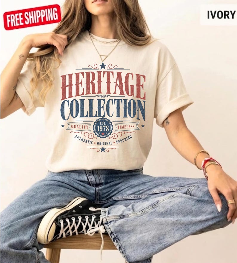 May include: Cream-coloured t-shirt featuring the text "HERITAGE COLLECTION" in red and blue. The shirt includes the text "EST. 1978" and other details. The model is wearing blue jeans and black trainers.