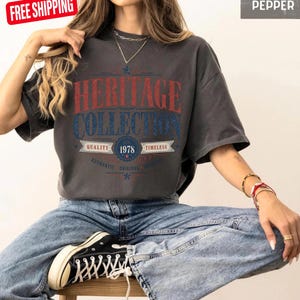 May include: A dark grey t-shirt with the text "HERITAGE COLLECTION" in red and blue. The shirt also features the year "1978" and the words "QUALITY" and "TIMELESS". The model is wearing blue jeans and black trainers.