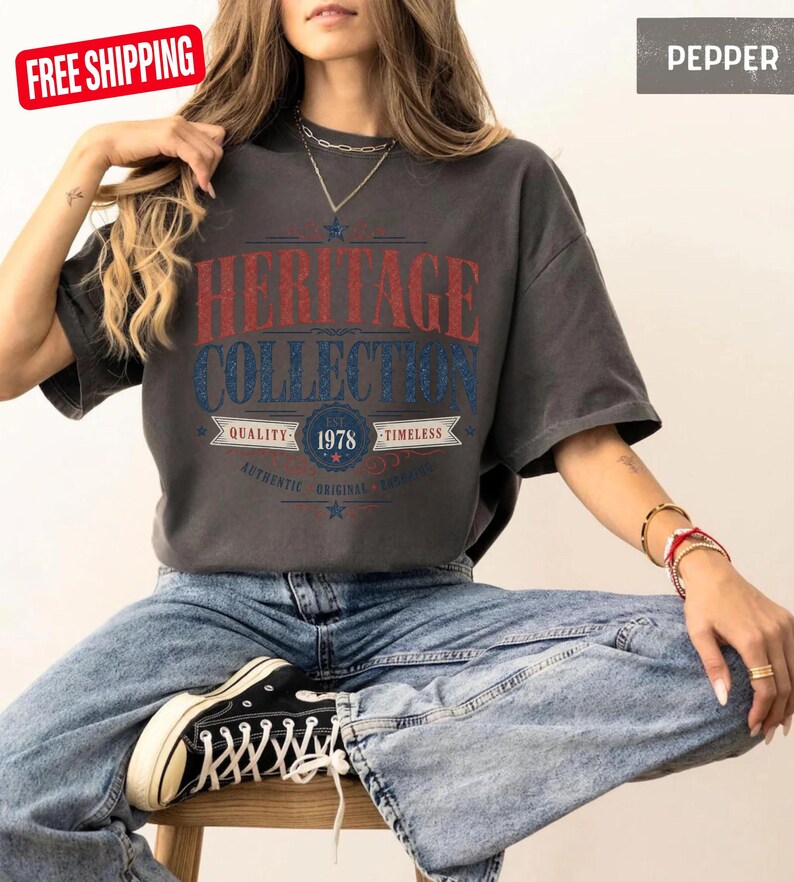 May include: A dark grey t-shirt with the text "HERITAGE COLLECTION" in red and blue. The shirt also features the year "1978" and the words "QUALITY" and "TIMELESS". The model is wearing blue jeans and black trainers.