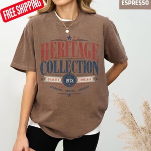 May include: Brown t-shirt with the text "HERITAGE COLLECTION" in red and blue. The shirt also features the text "EST. 1978" and the words "QUALITY", "TIMELESS", "AUTHENTIC", "ORIGINAL", and "ENDURING". The shirt is a solid brown colour.