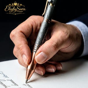 May include: A detailed shot of a pen with a patterned metal barrel and copper accents. The pen is held by a person writing on white paper. The pen's design includes a clip and a pointed tip. The Elegby Sara logo is visible in the upper left corner.