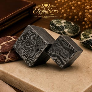 May include: Two dark gray metal cubes featuring a distinct, swirling Damascus steel pattern. The cubes are positioned at an angle, showcasing their geometric shape and layered texture. The logo ElegbySara Jewelry & Accessories is visible above.