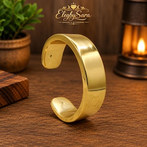May include: A polished gold-tone cuff bracelet with a smooth, wide band and an open design. The bracelet features a reflective metallic surface and rounded ends, showcasing a simple, continuous circular shape without any additional embellishments.