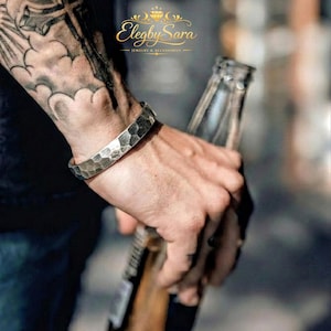 May include: A silver hammered cuff bracelet. The bracelet has a textured, geometric pattern. The image also shows a person holding a bottle. The brand name "Elegby Sara Jewelry & Accessories" is visible in the top right corner.