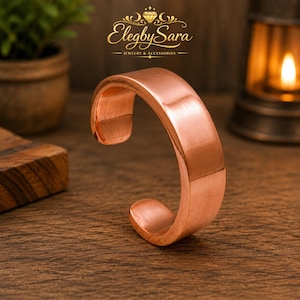 May include: A solid copper cuff bracelet with a smooth, polished finish and a wide, open-ended band design. The metal surface reflects light, showcasing the warm, metallic reddish-brown color of the copper material in a simple, circular shape.
