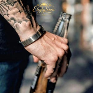 May include: A silver-colored cuff bracelet with a brushed metal finish. The bracelet is worn on a wrist. The image also shows a bottle. The jewelry is from Elegby Sara.