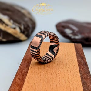 May include: A copper-toned ring with a geometric black stripe design. The ring is displayed on a wooden surface. The ring has a flat top and a patterned band. The ring is a piece of jewelry.