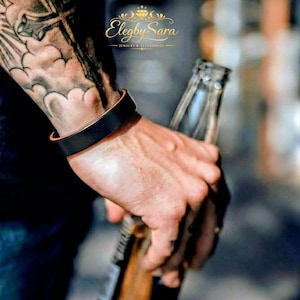 May include: A black leather bracelet with a copper-colored interior is worn on a wrist. The bracelet is the focal point, with the brand name "Elegby Sara Jewelry & Accessories" visible in the background. The person is holding a bottle.