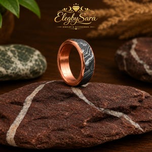 May include: A ring featuring a copper-colored inner band and a textured, dark gray outer band with a wavy, layered pattern. The logo ElegbySara Jewelry & Accessories is visible at the top center above the ring, which rests on a brown rock.