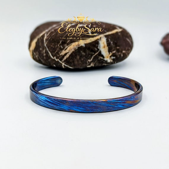Timascus (titanium Damascus) is one of the rarest, most visually spectacular metals on earth — the blue flame pattern on this hand-forged cuff is genuinely one-of-a-kind and looks like wearable dark fantasy art.