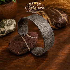 Peut inclure: Un bracelet manchette large en métal gris foncé présentant un motif circulaire répétitif sur toute sa surface. Le bracelet possède un design ouvert et une finition texturée. Le logo ElegbySara Jewelry & Accessories est visible au-dessus.