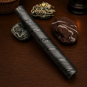 May include: A cylindrical rod featuring a dark gray and black Damascus steel pattern with wavy, wood-grain-like layers. The rod is positioned diagonally across the frame, displaying a polished metallic surface with intricate, flowing lines throughout.