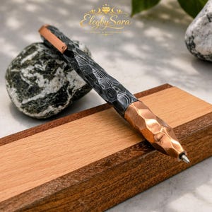 May include: A ballpoint pen featuring a dark, textured Damascus steel barrel with a wavy pattern, paired with a faceted, metallic copper-colored grip and clip. The brand name Elegby Sara is visible in gold text above the pen.