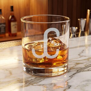 Puede incluir: Un vaso de cristal transparente con la letra C grabada y esmerilada en la parte frontal. El vaso contiene un líquido de color ámbar y varios cubitos de hielo transparentes, con una base pesada y paredes rectas para un recipiente clásico.