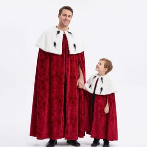Pode incluir: Duas capas a condizer até ao chão feitas de veludo vermelho profundo com um padrão floral subtil. Cada capa apresenta uma gola de pele sintética branca adornada com pequenos detalhes de penas pretas na frente, presa com um fecho prateado.